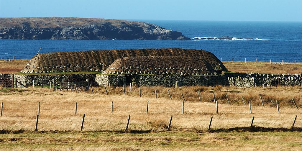 Two low thatched buildings are shown against a backdrop of cliffs, sea and a blue sky.
